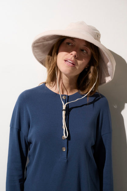 Femme portant un chapeau anti-UV beige UPF 50+ et un haut bleu, idéal pour la protection solaire à l'extérieur.