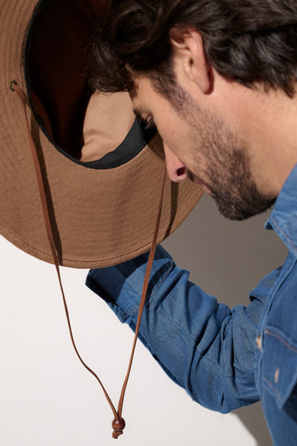 "Homme ajustant un chapeau anti-UV marron UPF50+, idéal pour la protection solaire lors d'activités en plein air."