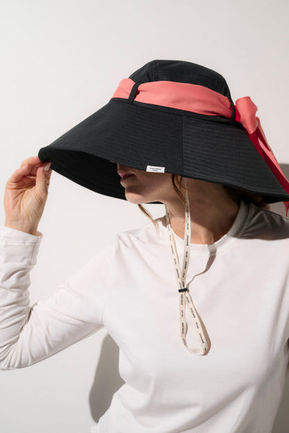 Chapeau anti-UV noir avec ruban rose, offrant protection solaire UPF50+, porté par une femme en vêtements légers.