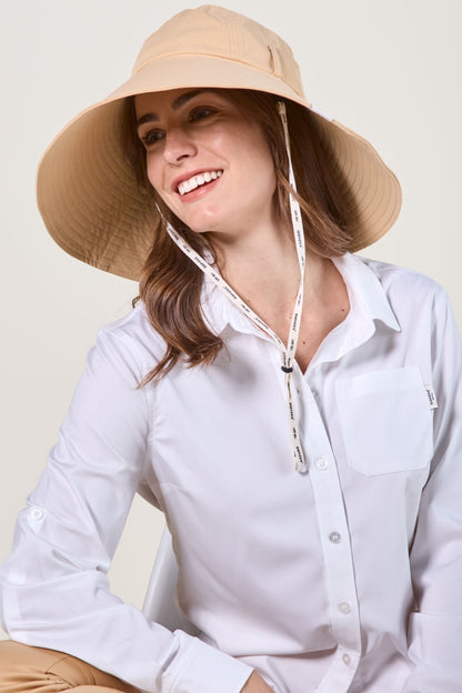 Femme souriante portant un chapeau anti-UV beige et une chemise blanche, idéale pour une protection solaire.